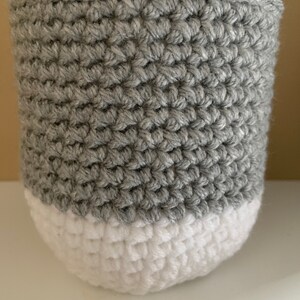 May include: A gray and white crocheted basket with a round shape. The basket is made of a soft, textured yarn.