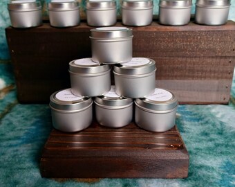 May include: A collection of small, silver-colored metal tins with white labels, arranged on dark wooden display stands. The tins are stacked in a pyramid shape, with some arranged on a higher level. The labels likely indicate the contents of the tins.