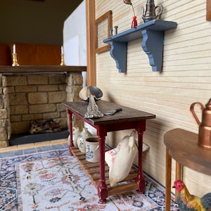 May include: A miniature dollhouse room with a wooden table, a red and white rug, a rooster figurine, and a shelf with a silver teapot and a red vase with a single red flower.