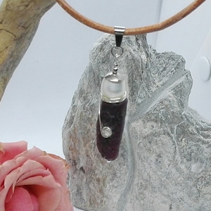 Puede incluir: Un collar con un colgante de plata, una piedra roja oscura y un cristal transparente en un cordón de cuero marrón.