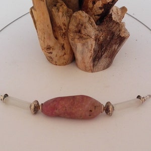 May include: A pink and white necklace with a large, irregularly shaped pink stone pendant. The pendant is surrounded by two silver beads and two white beads.