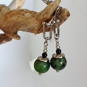 Könnte beinhalten: Ein Paar silberne Ohrclips mit grünen und schwarzen Perlen. Die grünen Perlen sind rund und haben ein geflecktes Muster. Die schwarzen Perlen sind klein und rund.