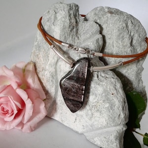 Peut inclure: Un collier en cuir marron avec un pendentif argenté orné d'une pierre gris foncé irisée. Le collier est présenté sur un rocher gris avec une rose rose au premier plan.