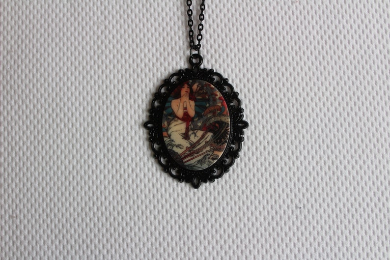 May include: A black necklace with a round pendant featuring a vintage illustration of a woman in a white dress. The pendant has a black filigree design around the edge.