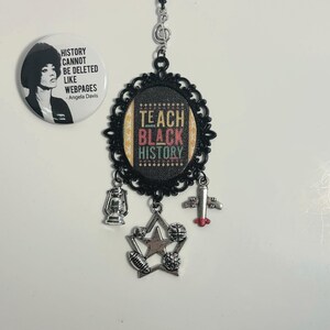 May include: A black necklace with a silver pendant featuring the words "Teach Black History" in colorful letters. The pendant is surrounded by a black filigree frame and has a silver star charm with other charms hanging from it.