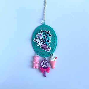 May include: Teal and white necklace with a charm of a woman holding a lollipop. The charm is surrounded by a teal filigree design. The necklace also has three charms hanging from it: a pink gummy bear, a purple lollipop, and a pink and white ice cream cone.