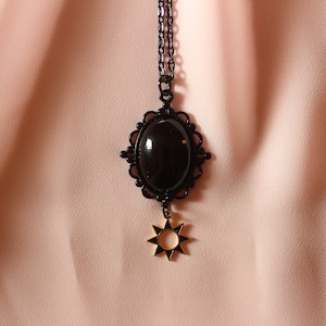 May include: A black necklace with a large oval pendant featuring a black gemstone surrounded by a black filigree design. A gold star charm hangs from the bottom of the pendant.