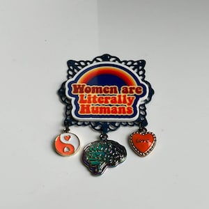 Puede incluir: Un pin de esmalte azul y blanco con un arcoíris y el texto "Women are Literally Humans". El pin tiene tres abalorios colgantes: un símbolo yin yang, un cerebro y un corazón con la palabra "Love" en él.