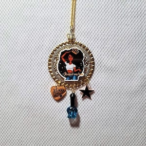 Op de afbeelding: Gouden ketting met een ronde hanger met een cartoonillustratie van een zwarte vrouw met een kroon en de tekst "Respect my hustle". De hanger is omgeven door een decoratief gouden frame. De ketting heeft ook een hartvormige hanger met de tekst "slay like me", een zwarte sterhanger en een blauwe nagellakhanger.