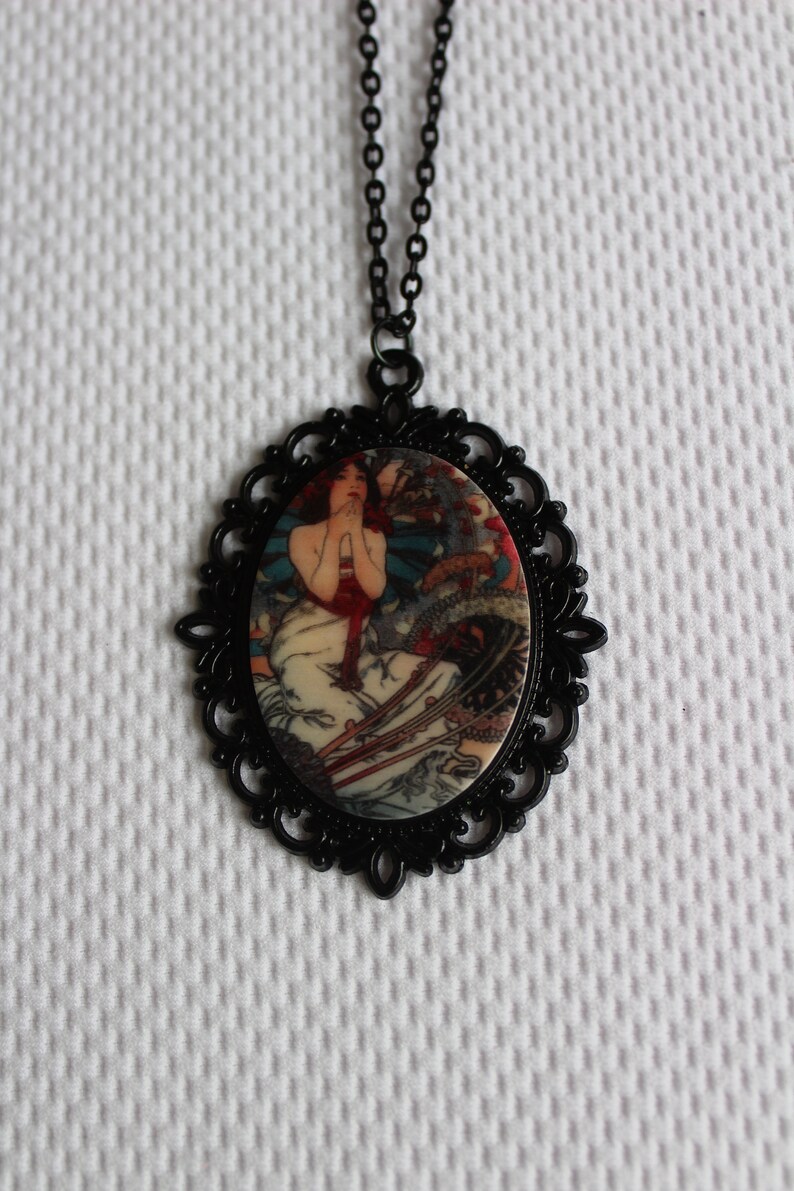 May include: A black chain necklace with a cameo pendant featuring a vintage illustration of a woman in a white dress with red accents. The cameo is surrounded by a black filigree frame.