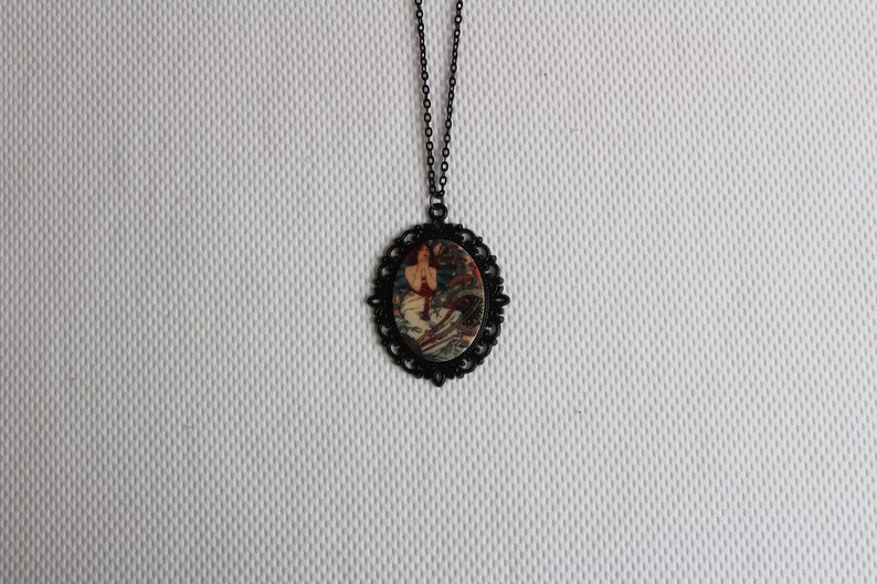 May include: A black chain necklace with a round pendant featuring a vintage illustration of a woman in a red dress. The pendant has a black filigree border.