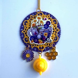 Op de afbeelding: Een paarse en gouden ketting met een theekop-bedel gevuld met bloemen, een madeliefje-bedel, een honingraat-bedel en een gele theepot-bedel.