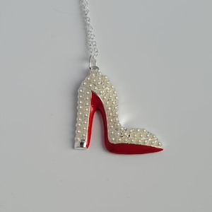Könnte beinhalten: Eine silberne Halskette mit einem roten und weißen High-Heel-Schuh-Anhänger. Der Schuh ist mit weißen Perlen besetzt.
