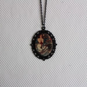 May include: A black chain necklace with a round pendant featuring a vintage illustration of a woman in a red dress. The pendant has a black filigree design around the edge.