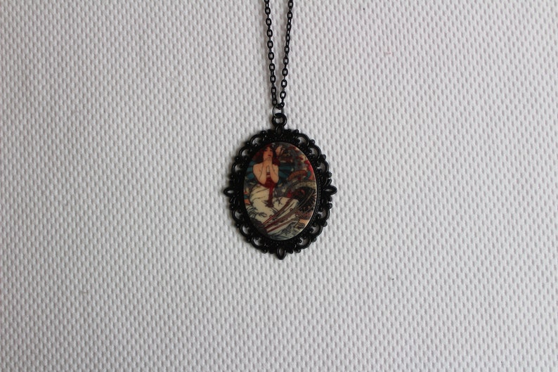 May include: A black chain necklace with a round pendant featuring a vintage illustration of a woman in a red dress. The pendant has a black filigree design around the edge.