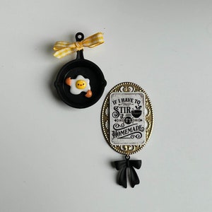 May include: A gold-colored metal charm with a white background and black text that reads "If I have to stir it's homemade". The charm has a black bow and is attached to a black frying pan with a yellow egg inside.