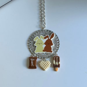 May include: A silver chain necklace with a round pendant featuring two cartoon bunnies, one brown and one white. The pendant is surrounded by a decorative silver filigree design. Hanging from the pendant are three charms: a brown chocolate bar, a white waffle heart, and a brown and white popsicle.