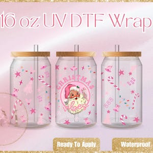 Puede incluir: Tres vasos de vidrio transparente con forma de lata con tapas de bambú, cada uno decorado con gráficos rosas con temática navideña. El vaso central presenta un diseño de Papá Noel con el texto "Christmas Vibes". El texto "16 oz UV DTF Wrap" está en la parte superior.