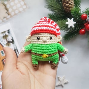 Puede incluir: Un pequeño adorno de elfo de crochet con un gorro a rayas rojas y blancas, cuerpo verde y zapatos marrones. El elfo tiene una cara amigable con ojos negros y un cinturón marrón.