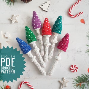 Puede incluir: Un conjunto de seis adornos de setas de ganchillo en varios colores, incluyendo rojo, verde, azul, morado y rosa. Cada seta tiene puntos blancos y un tallo blanco. Las setas están dispuestas sobre una superficie blanca con una estrella, un bastón de caramelo y otras decoraciones navideñas.