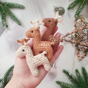 May include: Three crocheted reindeer ornaments in brown and white. The reindeer are standing on a hand.