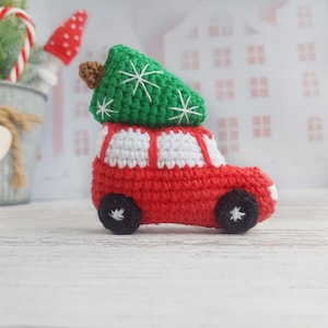Puede incluir: Un coche de juguete rojo de crochet con detalles blancos que lleva un árbol de Navidad verde de crochet en la parte superior. El coche tiene ruedas negras y detalles de copo de nieve blancos.