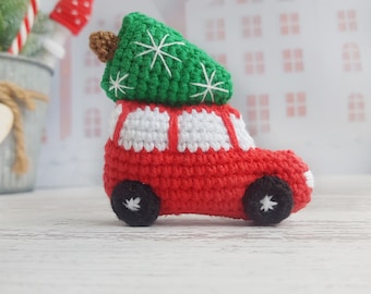 Peut inclure: Une petite voiture rouge en crochet avec des accents blancs transportant un sapin de Noël vert en crochet sur le dessus. La voiture a des roues noires et des détails de flocons de neige blancs.