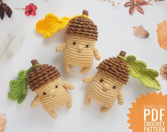 May include: Three crocheted acorn amigurumi toys with smiling faces. Each acorn has a crocheted leaf attached. The acorns are brown and beige, and the leaves are green and yellow. The image includes the text "ENGLISH PDF CROCHET PATTERN".