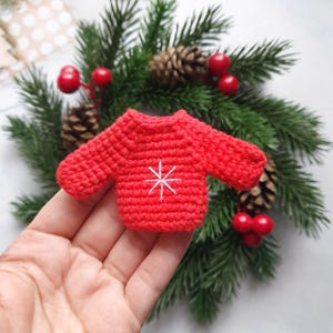 Könnte beinhalten: Ein kleiner roter Häkelpullover mit einem weißen Schneeflockenmuster. Der Pullover liegt auf einer Hand vor einem grünen Weihnachtsbaum.