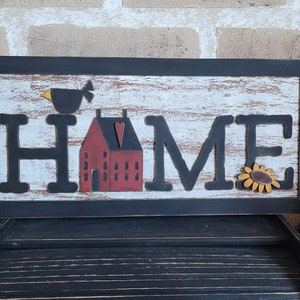 Może przedstawiać: Drewniany znak z napisem "HOME" czarnymi literami. Znak jest ozdobiony małym czerwonym domkiem, czarnym ptakiem i żółtym słonecznikiem.