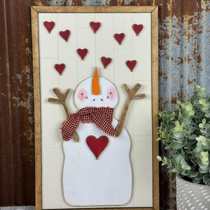 Op de afbeelding: Houten wandkunst met een sneeuwpop. De sneeuwpop heeft een rood hart, een geruite sjaal en een oranje wortelneus. Rode harten staan boven de sneeuwpop. De tekst "Pickitz Crafts" staat bovenaan.