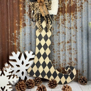 May include: A black and white checkered elf boot stocking with a burlap bow and a tag that says "Believe". The stocking is decorated with bells and a white fur cuff. The stocking is sitting on a white wooden surface with pine cones and a white snowflake.