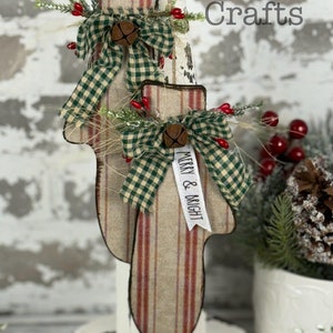 May include: Two decorative mitten-shaped ornaments. The mittens are made of fabric with red stripes and a plaid top. Each mitten has a green plaid bow, a small bell, and sprigs of greenery with red berries. The text "Merry & Bright" is on a white ribbon.