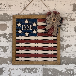 May include: A wooden framed American flag wall decor with red, white, and blue stripes and a blue field with white stars. The flag has the year 1776 in the center of the blue field. A red, white, and blue fabric bow is attached to the top of the frame.