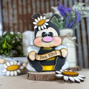 May include: Wooden bee figurine with black and yellow stripes, holding a banner that says "Bee Kind". The bee has a pink nose and large eyes. The figurine is decorated with white daisies.