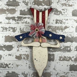 May include: A wooden patriotic gnome with a red, white, and blue hat, a white beard, and a blue hat with white stars. The gnome is hanging from a wire.