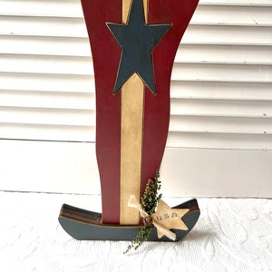 Puede incluir: Una bota de madera de temática patriótica pintada en rojo, blanco y azul. Una estrella azul está pintada en la parte delantera de la bota. La bota tiene un pequeño letrero que dice "USA" en él.