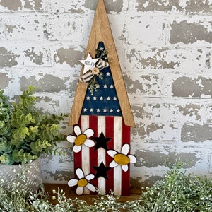 May include: Wooden patriotic-themed triangle-shaped sign with a red, white, and blue American flag design. The sign features white daisies with yellow centers and black stars. The sign has the text "U.S.A." on the top.