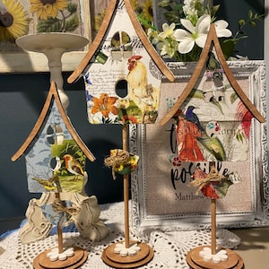 May include: Three wooden birdhouse figurines decorated with colorful paper and floral patterns. Each birdhouse is on a wooden stand with a white flower design. The birdhouses are arranged on a white crocheted doily.