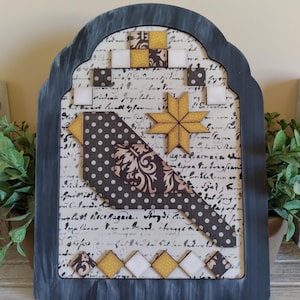 May include: A decorative wooden plaque with a dark gray frame. The artwork features a bird design with a polka dot pattern and a star, set against a backdrop of handwritten script. Accents include yellow and white squares.