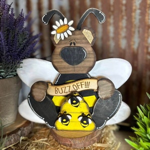 May include: Wooden bee-themed figurine with a brown bear head, white wings, and a yellow and black bee body. The bear is holding a banner that says "Buzz Off!!" and is sitting on a wooden base.