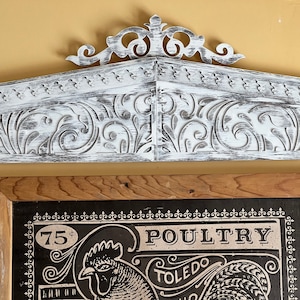Puede incluir: Un letrero de madera de estilo vintage con un acabado de pintura blanca desgastada. El letrero presenta una ilustración en blanco y negro de un gallo con el texto "75¢ POULTRY TOLEDO" encima.