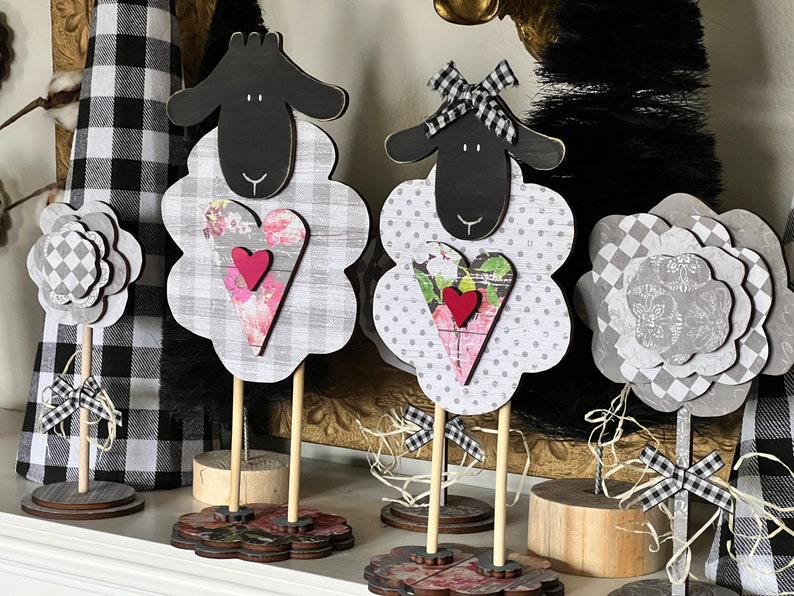 May include: Three black and white sheep figurines with floral patterned hearts and a gray and white checkered flower figurine. The figurines are on wooden stands with black and white checkered bows.