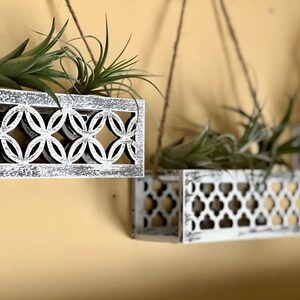 May include: Three white wooden hanging planters with a geometric design. Each planter is filled with green air plants.