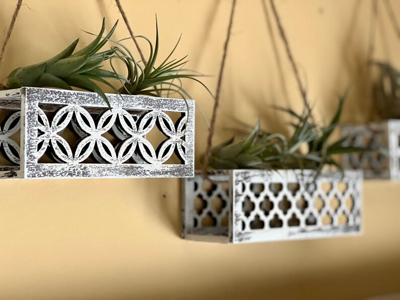 May include: Three white wooden hanging planters with a geometric design. Each planter is filled with green air plants.