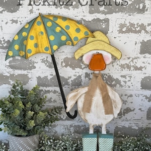 May include: A wooden duck figurine wearing a yellow hat and a scarf, holding a yellow and blue polka dot umbrella. The duck is wearing blue and white chevron rain boots. The background is a white brick wall.