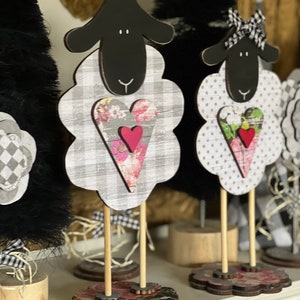 May include: Three black and white sheep figurines with hearts on their chests. The sheep are made of wood and have a rustic, farmhouse style. They are standing on wooden bases with floral patterns.