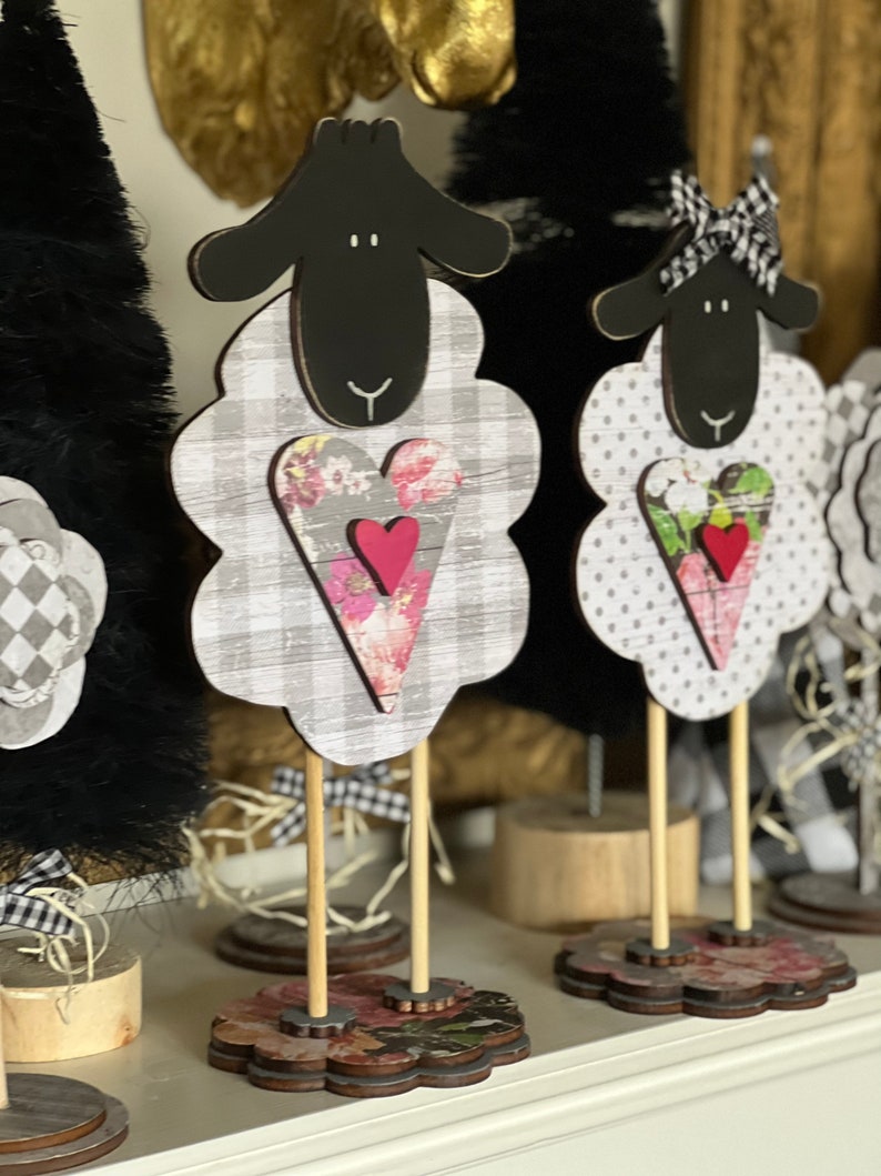 May include: Three black and white sheep figurines with hearts on their chests. The sheep are made of wood and have a rustic, farmhouse style. They are standing on wooden bases with floral patterns.