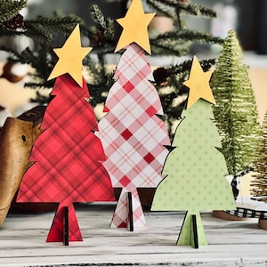 May include: Three wooden Christmas tree ornaments with a red plaid, pink and white plaid, and green polka dot design. Each tree has a gold star on top.