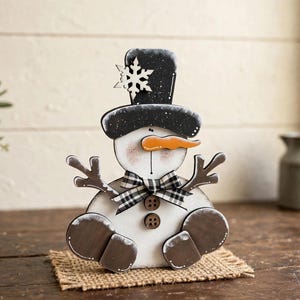 Op de afbeelding: Een houten sneeuwpopfiguur met een zwarte hoge hoed voorzien van een witte sneeuwvlok, een oranje wortelneus en een zwart-wit geblokte strik. De sneeuwpop heeft twee bruine knopen op de borst en donkerbruine takachtige armen.
